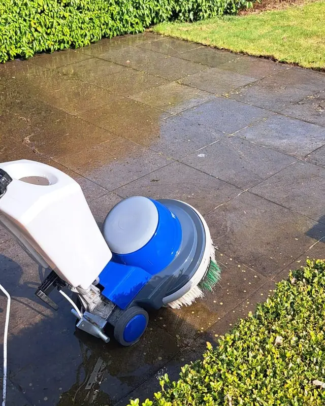 Borstelmachine met groene borstel op grijze tegels naast een haag